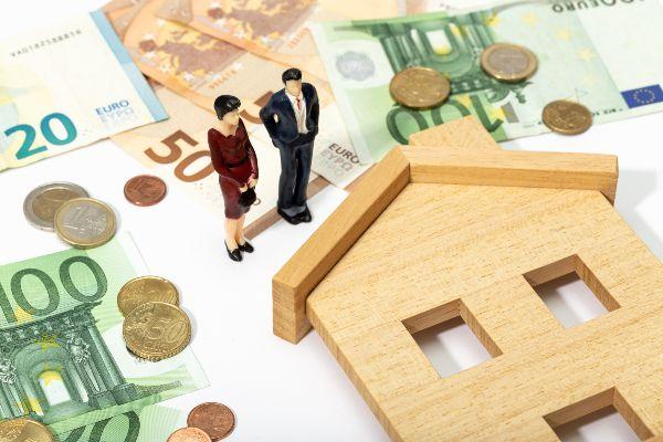 Como vender una casa; imagen de una casa de madera rodeada de dinero y dos pequeños muñecos como símbolo de la venta de una propiedad.
