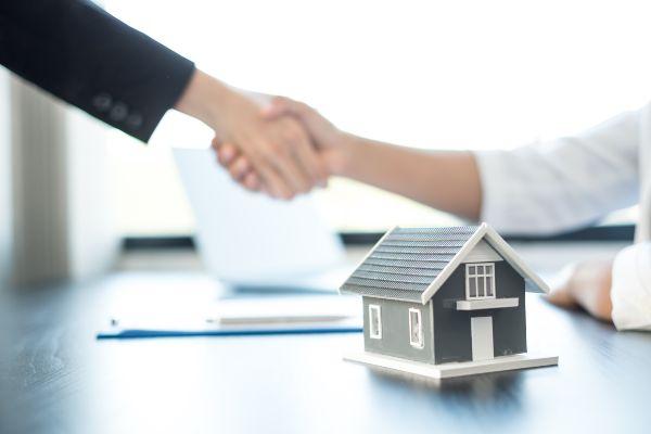 Comprar casa; imagen de dos personas cerrando un trato de la compra de una casa con un apretón de manos de fondo.
