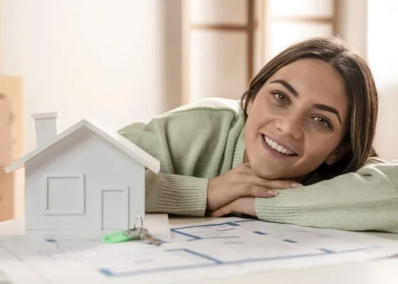 Mujer sonriente sobre una mesa donde se ve una maqueta de casa y papeles, transmitiendo la tranquilidad de avanzar en trámites relacionados con un Credito Infonavit.